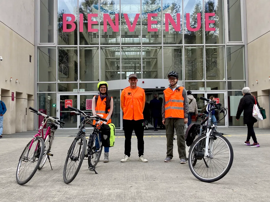 Trois cyclistes en gilet orange avec leurs vélos devant l'entrée d'un bâtiment de l'Université de Genève